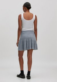 Vrouw met een bun, gekleed in een witte mouwloze top, een grijze textuur flared rok, zwarte sokken en zwarte instappers, staand met de rug naar de kijker.