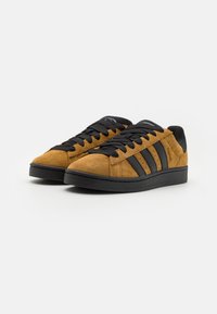 Brown suede sneakers with black rubber sole, black laces, and three black stripes on each side. Rounded toe and textured surface.