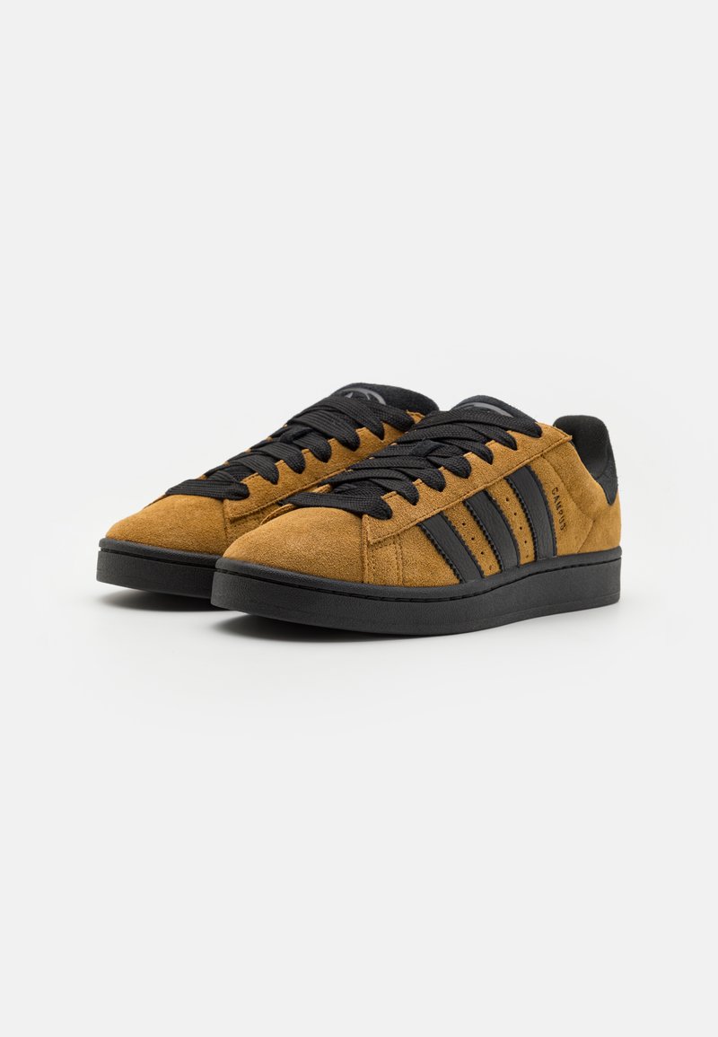 Zapatillas de ante marrón con suela de goma negra, cordones negros y tres rayas negras a cada lado. Punta redondeada y superficie texturizada.