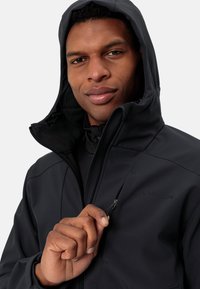 Black softshell jacket with a zippered front and hood, featuring a textured interior and logo detail on the chest. Close-up of hand adjusting zipper.