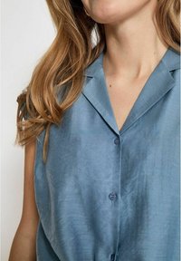 Camicetta a maniche corte di colore azzurro chiaro realizzata in un tessuto liscio, con colletto a punta e chiusura frontale con bottoni.