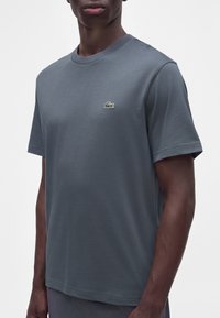 Homme portant un t-shirt gris foncé uni à manches courtes avec un petit logo crocodile vert sur le côté gauche de la poitrine.