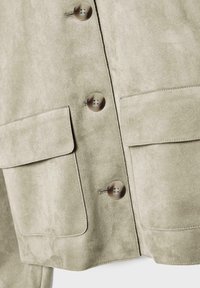 Veste en daim vert clair avec deux grandes poches frontales et des boutons ronds marron, caractérisée par une texture douce et des lignes épurées.