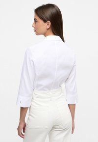 Camicia bianca con colletto, maniche a tre quarti e tessuto strutturato, con cucitura sul retro visibile, abbinata a pantaloni bianchi a vita alta.