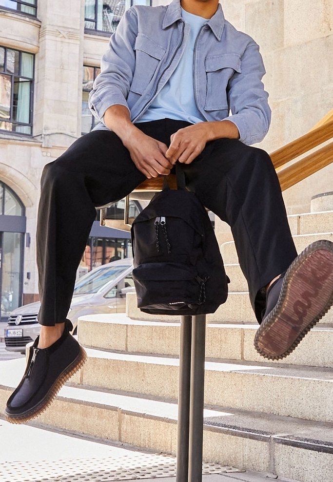 Personne portant une veste légère et un pantalon noir assise sur des escaliers, tenant un sac à dos noir, portant des chaussures noires à semelles épaisses.