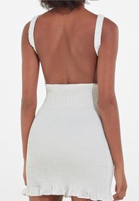Bílá šaty s nařasením, bez zad, širokými ramínky, strukturovanou tkaninou a volánovým lemem. Hladký povrch s jemným elastickým efektem.