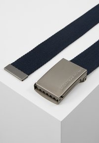 Ceinture tissée bleu marine avec une boucle en métal au fini brossé et du texte gravé. La boucle a un bord dentelé pour un maintien sécurisé.