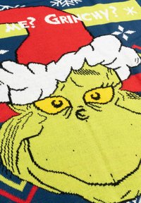 Coperta in maglia con il volto del Grinch in verde e occhi gialli, che indossa un cappello di Babbo Natale rosso con bordi bianchi, su uno sfondo blu.
