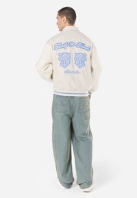 Beige bomberjakke med hvite ribbede mansjetter, stort blått broderi med løver og tekst, brukt sammen med baggy lyseblå jeans.
