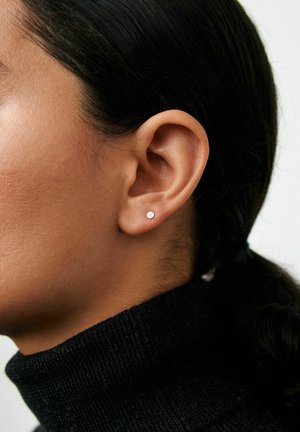 Detail osoby s malým okrúhlym náušnicovým gombíkom, oblečený v čiernom roláku.