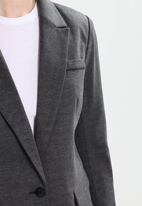 Blazer grigio su misura con rever a gatto, tessuto testurizzato, chiusura a un bottone e tasca sul petto, indossato sopra un top rosa chiaro.