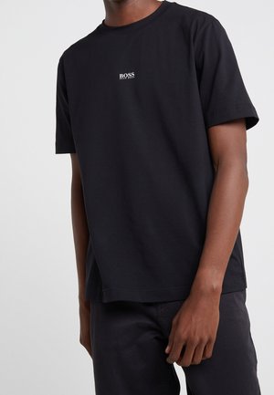 Muž má na sobě černé tričko s krátkým rukávem a malým bílým logem "BOSS Hugo Boss" umístěným uprostřed na hrudi.