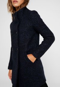 Cappotto in misto lana blu navy caratterizzato da un tessuto testurizzato, chiusura doppiopetto, due grandi bottoni, tasche laterali e colletto alto.