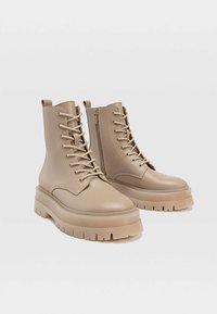 Botas de tobillo beige con suelas gruesas, cordones en la parte frontal, cremallera lateral, material sintético suave y punteras redondeadas. Detalle texturizado en la suela.