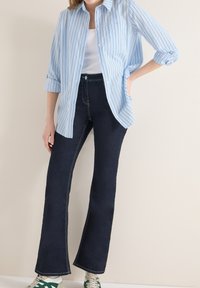 Femme portant un jean évasé bleu foncé, un débardeur blanc, une chemise à rayures bleues et blanches à manches retroussées, et des baskets blanches avec des rayures vertes.