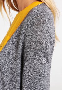 Grå texturerad cardigan som bärs över en gul skjorta, med en avslappnad passform och öppen framsida, samt en synlig ficka på skjortan.