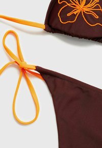 Rjav bikini zgornji del z oranžnimi naramnicami in vezenim cvetličnim vzorcem. Naramnice imajo obliko vozla za prilagodljiv fit. Gladka tekstura tkanine.