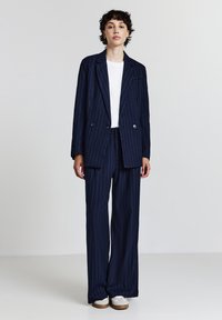 Completo in tessuto rigato navy caratterizzato da un blazer doppiopetto con rever a punta e pantaloni a gamba larga. Indossato sopra una maglietta bianca.