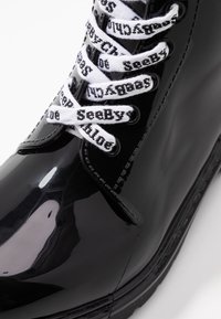 Bottines en cuir verni noir avec des lacets blancs à logo, bout arrondi, texture lisse et surpiqûres visibles le long du bord.