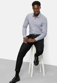 Himmelblaues Hemd mit kleinen dunkelblauen Pünktchen, kombiniert mit schwarzen Slim-Fit-Hosen und schwarzen eleganten Schuhen, sitzt auf einem weißen Hocker.