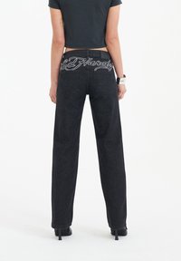 Black high-waisted jeans with a straight leg, featuring white embroidered "Ed Hardy" logo on the back waistband. Smooth denim texture.
