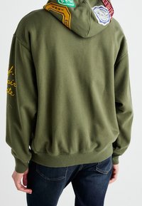 Hoodie vert olive avec une coupe décontractée, dotée d'une capuche à cordon et de patchs décoratifs jaunes, verts et rouges au dos.