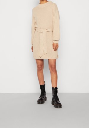 Osoba ubrana w beżową dzianą sukienkę z przewiązanym paskiem oraz czarne sznurowane wojskowe buty, stojąca na tle gładkiej ściany.