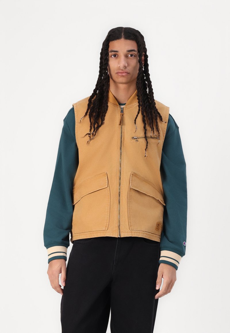 Giacca senza maniche color sabbia con chiusura a zip e due grandi tasche frontali, abbinata a una camicia a maniche lunghe teal con polsini a righe.