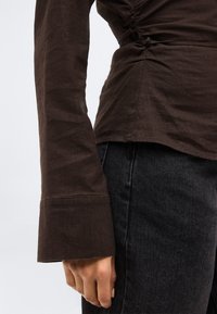 Πλάγια όψη ενός ατόμου που φοράει καφέ εφαρμοστό μακρυμάνικο τοπ και μαύρο τζιν ψηλόμεσο, με το αριστερό χέρι χαλαρό δίπλα στο σώμα.