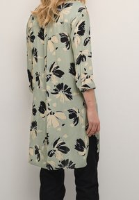 Tunique florale en tissu vert clair avec des motifs de fleurs noirs et beiges. Présente une coupe ample, des manches trois quarts et des fentes sur les côtés.