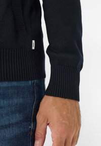 Marineblauwe gebreide trui met ribgebreide manchetten en zoom, voorzien van een klein logo-label aan de zijkant, gecombineerd met denim jeans. Close-up van een hand die de stof vasthoudt.