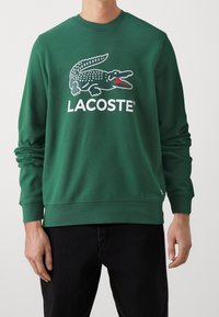 Zelená bavlněná mikina s velkým bílým logem krokodýla Lacoste s červenými akcenty, lemovaná ribem na rukávech a spodním lemu.