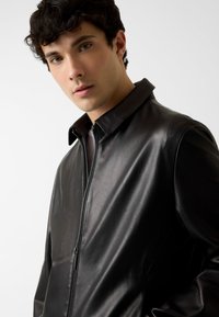 Zwarte leren jas met een gladde textuur, voorzien van een hoge kraag en een voorrits. Het ontwerp is minimalistisch en op maat gemaakt.