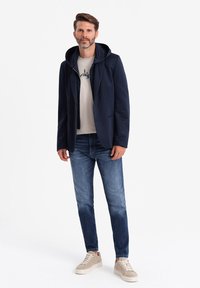Marine blazer med hætte, strækstof og forlommer, parret med en lys grå t-shirt, mørke jeans og beige sneakers.