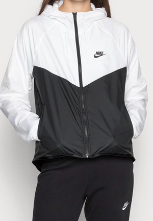 Kobieta ubrana w czarno-białą kurtkę Nike na zamek z kapturem, z rękami w kieszeniach, w połączeniu z czarnymi spodniami Nike.