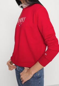 Červená mikina s kulatým výstřihem. Obsahuje bílé vyšívané logo "TOMMY JEANS". Hřejivé manžety a spodní lem, měkká tkanina, nošena s modrými džínami.