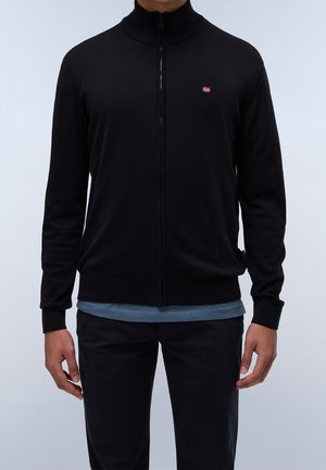 Mężczyzna ubrany w czarny sweter na zamek z małym logo w kształcie czerwono-biało-niebieskiej flagi na lewej piersi, pod nim niebieska koszula i czarne spodnie.