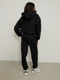 Juodas džemperis su kapišonu ir sportinės kelnės, pasižymintys laisvu siluetu, tekstūruotu audiniu ir erdviu kapišonu, derinami su baltais ir juodais sportiniais batais.