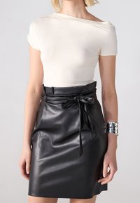 Vit av-axel topp i kombination med en åtsittande svart läder mini kjol med knytning i midjan och glänsande yta. Silverfärgad manschettarmband på handleden.