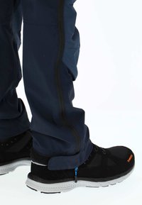 Granatowe wodoodporne spodnie z czarnym paskiem z boku, zestawione z czarnymi butami sportowymi z białą podeszwą i pomarańczowym logo.