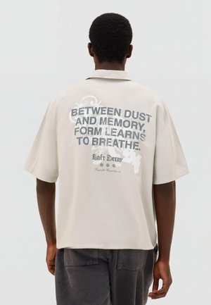 Mand med mørk hud og kort krøllet hår iført beige skjorte med teksten "Mellem støv og hukommelse lærer formen at ånde" på ryggen.