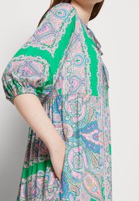 Zelené šaty s barevnými paisley vzory, vyrobené s nadýchanými rukávy a kapsami. Látka má hladkou strukturu a splývavou design.