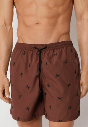 Mann trägt braune Badeshorts mit schwarzen Gecko-Motiven und schwarzem Kordelzug, zeigt Oberkörper und Oberschenkel.