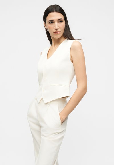 Γυναίκα με ίσια σκούρα μαλλιά, φοράει αμάνικη λευκή μπλούζα και ταιριαστό παντελόνι με ψηλή μέση, με τα χέρια στις τσέπες, κοιτάζει δεξιά.