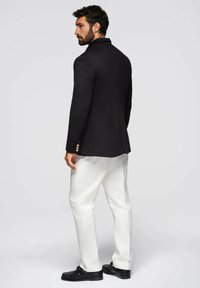 Schwarzer, taillierter Blazer mit Satinoberfläche, Ein-Knopf-Verschluss und goldenen Akzenten, kombiniert mit weißen Hosen und schwarzen Schuhen.