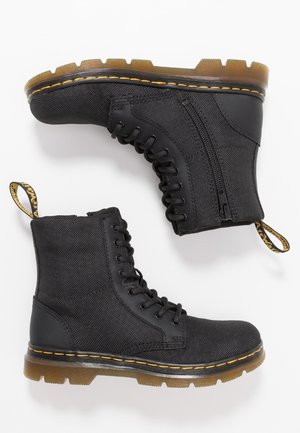 Bottines noires en tissu durable avec une finition texturée, dotées d'un laçage, d'une fermeture éclair sur le côté et de coutures jaunes sur une semelle épaisse.