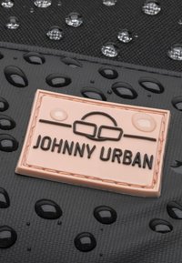 Čierna textúrovaná tkanina s kvapkami vody; obsahuje ružovú gumenú nášivku s nápisom "JOHNNY URBAN" s embosovaným písmom.