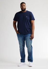 Námořnická modrá tričko s kulatým výstřihem a malým logem, kombinované s světlými modrými džíny a bílými teniskami. Volný střih, hladká textura.