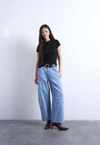 Νεαρή γυναίκα με μακριά σκούρα μαλλιά που φοράει μαύρο t-shirt, φαρδιά μπλε τζιν, μαύρη ζώνη και καφέ σκούρα μποτάκια μέχρι τον αστράγαλο, στέκεται σε τσιμεντένιο πάτωμα.