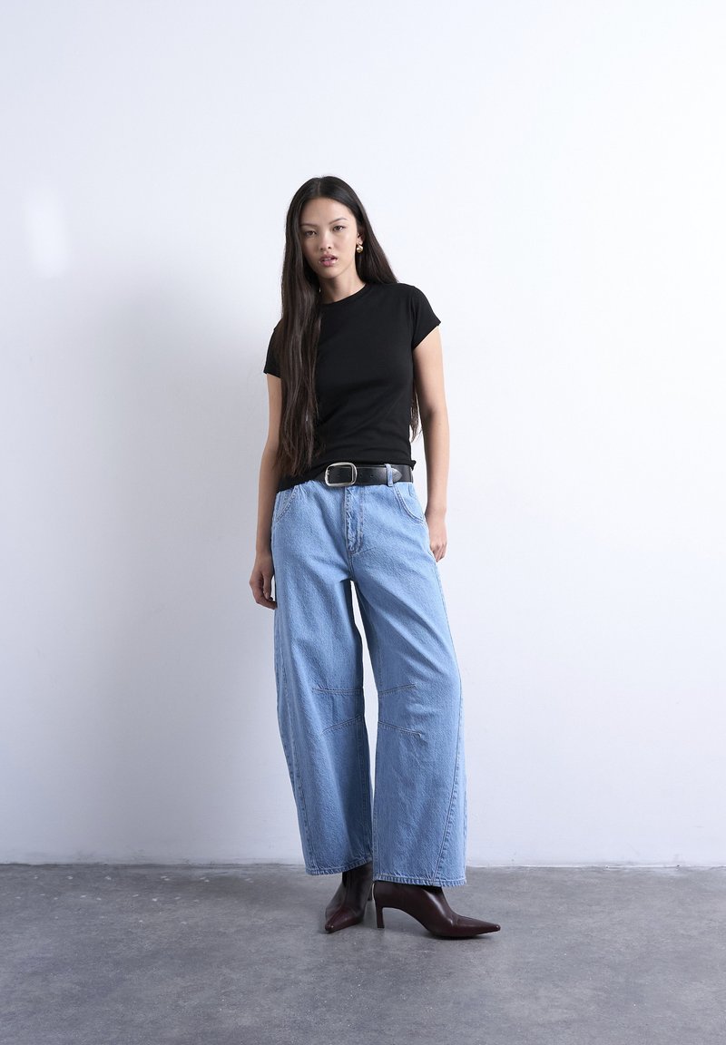 Νεαρή γυναίκα με μακριά σκούρα μαλλιά που φοράει μαύρο t-shirt, φαρδιά μπλε τζιν, μαύρη ζώνη και καφέ σκούρα μποτάκια μέχρι τον αστράγαλο, στέκεται σε τσιμεντένιο πάτωμα.